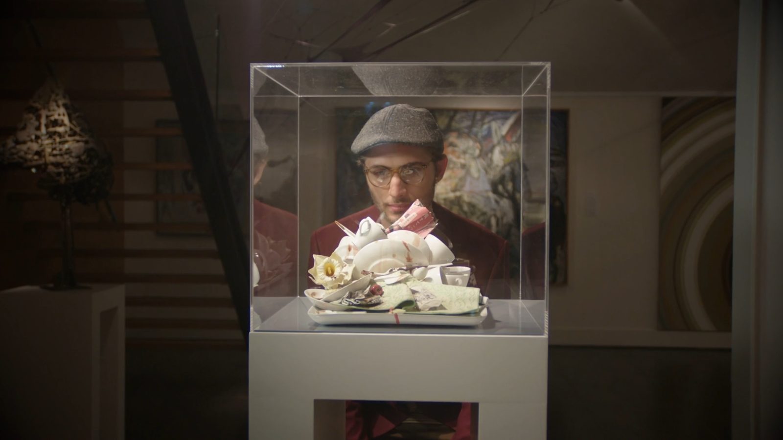 Fountain Avenue editing reel still: a man in glasses and a flat cap looking through a glass display case in a gallery