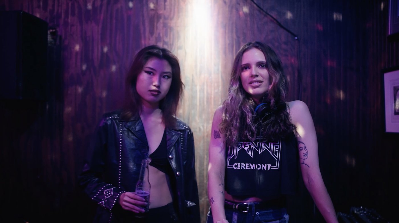 Q Train still: two women standing in a dim purple-lit club