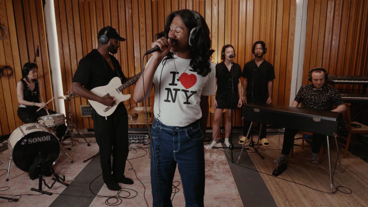 Live at Sheer Studios still: a singer in headphones and an I love NY shirt performing with a band in a wood-paneled studio